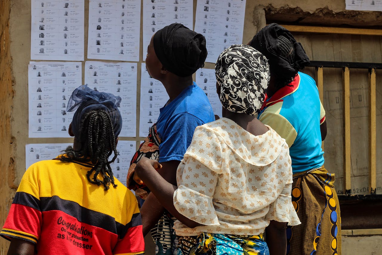 about-me-01 A group of women reading election lists posted on a wall outdoors during the day.