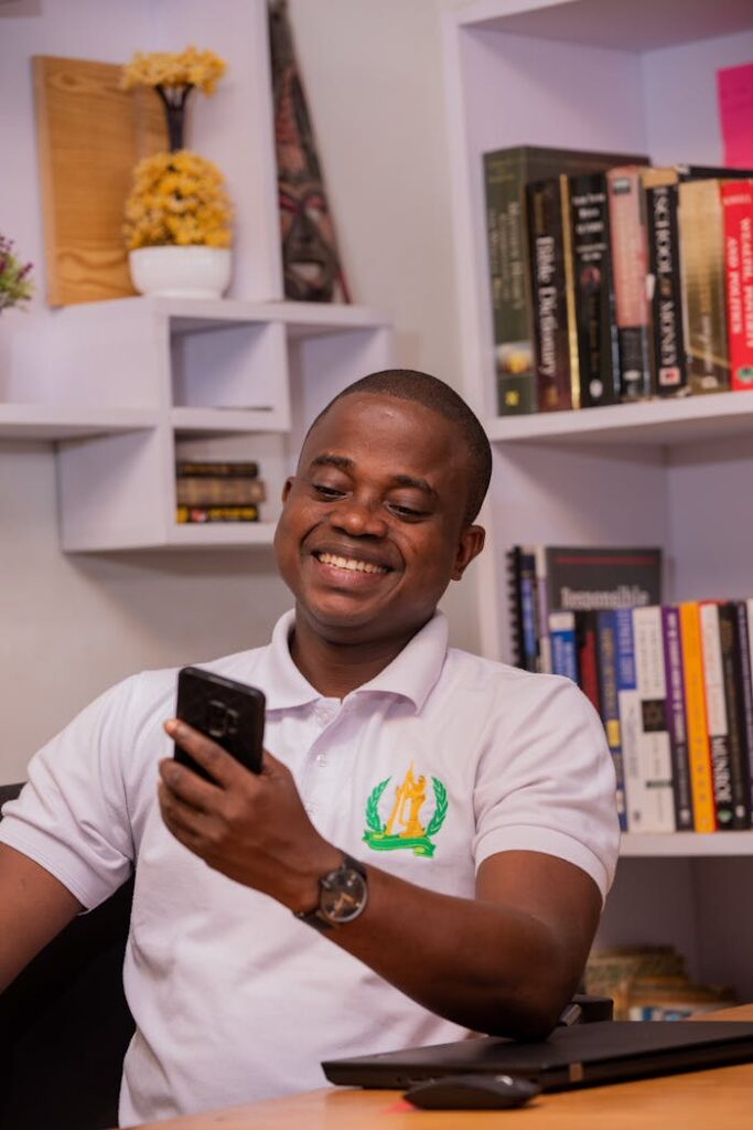 Happy man in a white polo shirt checking his smartphone while sitting at a desk in an office.