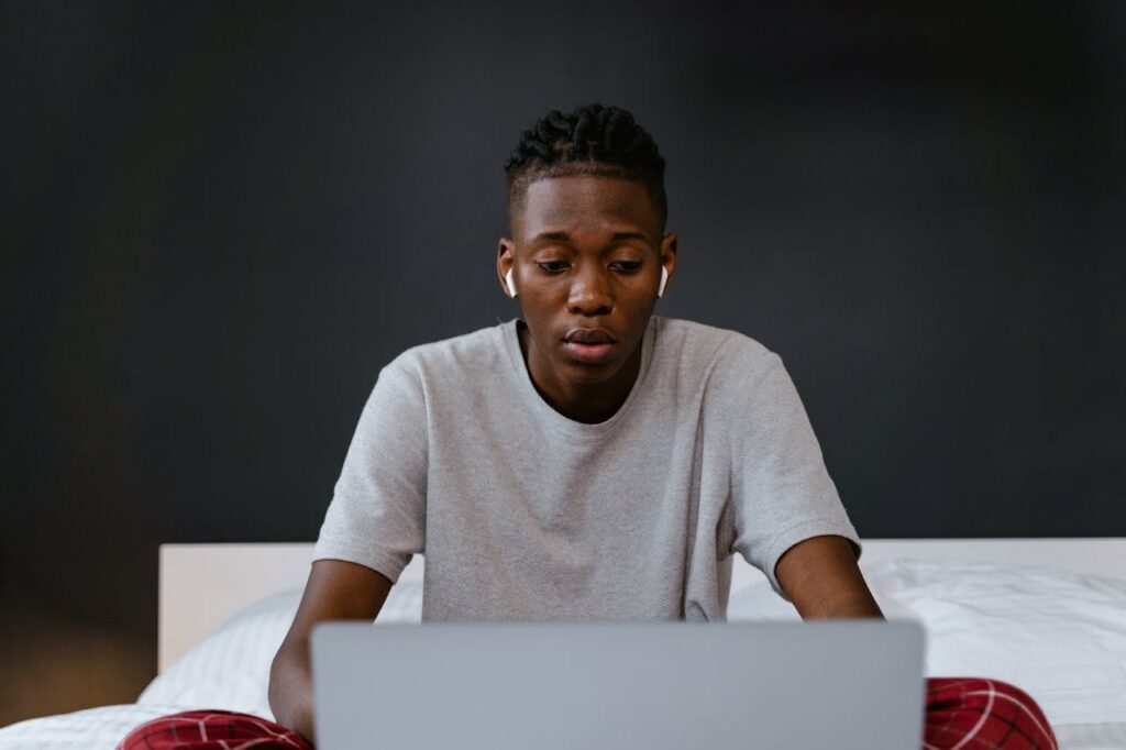 pexels photo 7606071 Young man focused on laptop screen while working from home, sitting on bed.